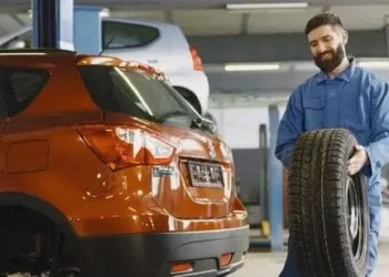Mehaničar u plavom kombinezonu drži auto gumu pored narandžastog vozila u radionici