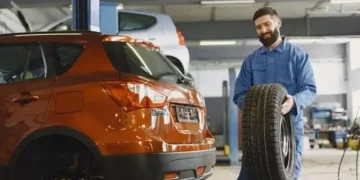 Mehaničar u plavom kombinezonu drži auto gumu pored narandžastog vozila u radionici